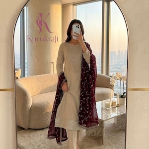 May include: A beige long-sleeved kurta set featuring a round neckline with intricate silver embroidery, paired with matching straight-leg trousers and a deep maroon sheer dupatta adorned with delicate white floral embroidery and a silver border.