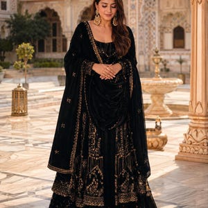 May include: A black floor-length lehenga choli featuring intricate gold embroidery throughout the skirt and bodice. The ensemble includes a matching black dupatta draped over the shoulders, adorned with gold detailing along the borders.