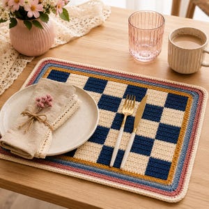 May include: A rectangular crochet placemat featuring a checkered pattern in navy blue and cream, bordered by thin stripes of mustard yellow, light blue, and dusty pink. The placemat is topped with a beige ceramic plate, a linen napkin, and gold cutlery.