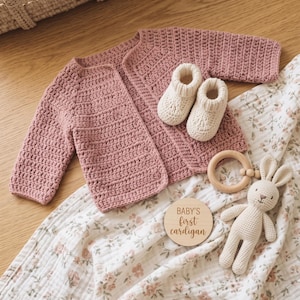 May include: A dusty rose crochet cardigan with long sleeves and an open front, paired with cream-colored knit booties, a wooden teething ring, a small crocheted rabbit toy, and a round wooden plaque inscribed with the text BABY'S first cardigan.