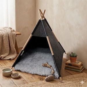 May include: A pet teepee constructed with light-colored wooden poles and dark gray felt panels. The interior features a plush, light gray faux fur mat. A small felt mouse toy with a rope tail sits on the floor in front of the open triangular entrance.