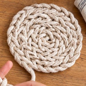 May include: A thick, off-white cotton rope being braided by hand. A large ball of the same rope sits to the left, and a finished circular, coiled rope mat lies to the right. The rope has a twisted texture and is made of natural fibre strands.