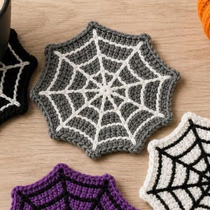 Könnte beinhalten: Ein Set aus vier gehäkelten Spinnennetz-Untersetzern in Schwarz, Grau, Weiß und Lila, jeweils mit einem kontrastierenden Netzmuster. Eine schwarze Tasse mit Kaffee steht auf dem schwarzen Untersetzer, neben einem kleinen gehäkelten Kürbis.