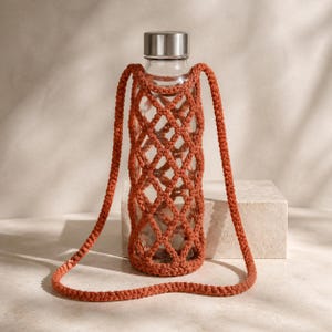 May include: A clear glass water bottle with a metallic silver screw-top lid, encased in a burnt orange macrame net holder featuring a diamond-shaped pattern and a long, matching cord strap for carrying the bottle over the shoulder or across the body.