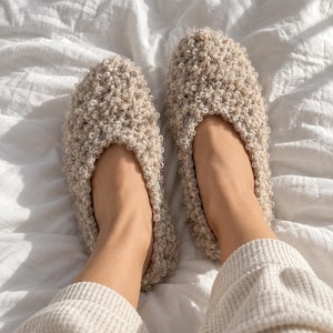 Op de afbeelding: Een paar beige slippers met een textuur van lussen garen. De slippers hebben een rond teenontwerp en een instapmodel, gemaakt van een dik, pluizig materiaal dat het gehele buitenoppervlak van het schoeisel bedekt.
