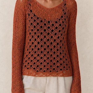 May include: A long-sleeved, rust-colored open-knit sweater featuring a complex, repeating geometric pattern. The sweater is layered over a solid black tank top and paired with light beige trousers, creating a layered look with visible texture.