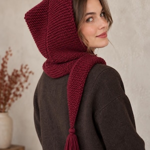 May include: A deep burgundy knitted hood with a garter stitch texture, featuring a long pointed tail that ends in a matching yarn tassel. The hood is designed to cover the head and wrap around the neck, providing a structured yet soft appearance.