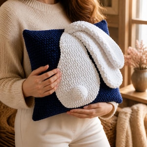 May include: A square navy blue crochet pillow featuring a large, plush white bunny face appliqué. The bunny design includes two long, soft white ears extending from the top and a small, round white tail centered on the dark blue textured surface.
