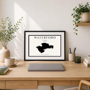 May include: A black framed wall art print featuring a cream background with a black silhouette map of Waterford, Ireland. The text reads WATERFORD, 52.2594°N 7.1103°W, and THE DÉISE, centered within a black rectangular border on the print.