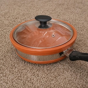 May include: An orange and silver cooking pan with a clear glass lid and a black handle. The pan has a round shape and is sitting on a carpeted surface. The lid has a black knob.
