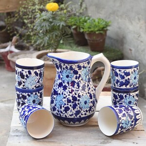 May include: A ceramic pitcher and matching cups set. The pitcher has a handle and a spout. The set is decorated with blue floral patterns on a white background. The cups are cylindrical and have a similar design. The set is placed on a wooden table.