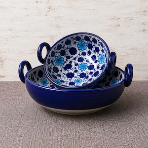 May include: Two blue ceramic bowls with handles, one nested inside the other. The interior of the bowls features a white background with a blue floral pattern. The exterior is a solid, deep blue.