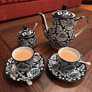 Pode incluir: Um conjunto de chá em cerâmica composto por um bule, um açucareiro com tampa e duas chávenas com pires, decorados com um padrão floral em azul escuro e branco. Cada chávena contém chá castanho claro e duas colheres de prata estão colocadas.