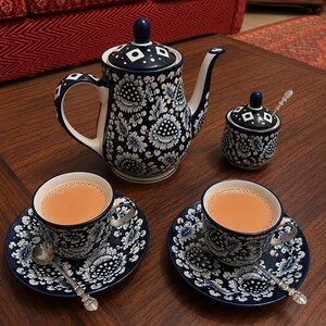 May include: A ceramic tea set featuring a dark blue and white floral pattern. The set includes a lidded teapot, a lidded sugar bowl with a small spoon, and two cups on matching saucers, each filled with light brown tea and accompanied by a silver spoon.