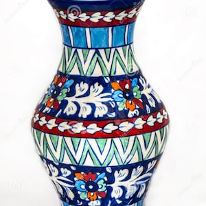 May include: A ceramic vase featuring a hand-painted design with bands of blue, red, and white. The pattern includes floral motifs, geometric zig-zag lines, and vertical stripes. The vase has a flared rim and a rounded body with a narrow neck.