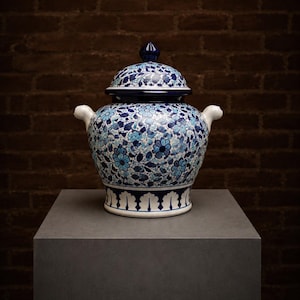 Op de afbeelding: Een keramieken gemberpot met een witte basis en ingewikkelde blauwe en lichtblauwe bloemmotieven. De pot is voorzien van een bijpassend deksel met een puntige knop, twee kleine handvatten aan de zijkanten en een decoratieve schulprand.