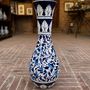Puede incluir: Un jarrón de cerámica alto con base blanca y patrones florales intrincados en azul oscuro y azul claro, junto con enredaderas. El cuello y la base están decorados con un motivo de hojas repetitivo y el borde es acampanado y brillante.