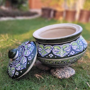 May include: A ceramic pot with a lid, featuring a dark green base and lid with a white rim. The pot is adorned with a repeating pattern of green and white floral designs. The lid is placed beside the pot on a bed of grass and rocks.