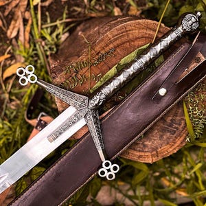 Op de afbeelding: Een zilverkleurig metalen zwaard met een ingewikkelde pareerstang met vier cirkelvormige uiteinden en een getextureerd handvat. Het zwaard is gecombineerd met een donkerbruine lederen schede met een zichtbare riem en een metalen knopsluiting.