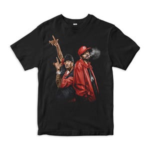 May include: A black short-sleeved t-shirt featuring a graphic print of two individuals. One individual wears a black beanie and red shirt, while the other wears a red baseball cap and red jacket with smoke visible near their face.