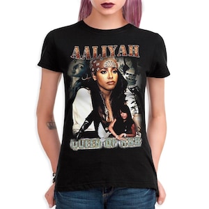 May include: A black short-sleeve t-shirt featuring a graphic print of the singer Aaliyah. The design includes the text AALIYAH at the top and QUEEN OF R&B at the bottom, with multiple photographic portraits of the artist and a small white dove.