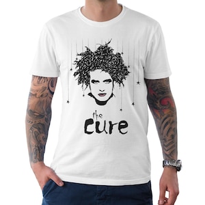 May include: A white short-sleeved t-shirt featuring a black graphic illustration of a person with messy hair and small stars hanging from strings above their head. The text "the CURE" is printed in black below the portrait in a stylized font.