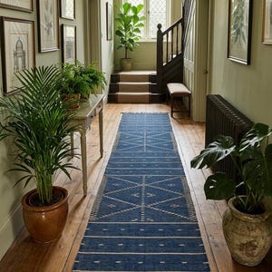 Puede incluir: Un pasillo con una alfombra azul con un diseño geométrico. La alfombra está sobre un suelo de madera, con plantas en macetas y obras de arte enmarcadas en las paredes. Una escalera es visible en el fondo.