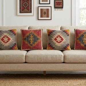 Puede incluir: Un sofá beige de tres cojines con cuatro cojines decorativos cuadrados con patrones geométricos en rojo, gris, amarillo y crema. El sofá tiene una tapicería de tela texturizada y patas de madera clara, ofreciendo una base neutra para los cojines.