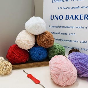 Op de afbeelding: Een verzameling meerkleurige fluwelen garenbollen in wit, crème, bruin, rood, blauw, groen, roze en paars, gerangschikt in een stapel. Een haaknaald met een rood handvat ligt op de voorgrond naast twee kleine gevlochten rotan bollen.