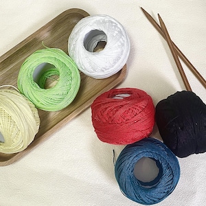 Peut inclure: Six pelotes de laine aux couleurs blanc, vert clair, jaune pâle, rouge, noir et bleu foncé, accompagnées de deux aiguilles à tricoter en bois. Trois pelotes sont sur un plateau en bois rectangulaire, les autres sont posées à côté.