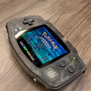 Könnte beinhalten: Eine durchscheinend graue Game Boy Advance Handheld-Spielkonsole mit einem Bildschirm, der den Titelbildschirm von Pokémon Emerald Version anzeigt, sowie einem Steuerkreuz, A- und B-Tasten und Start- und Select-Tasten auf der Vorderseite.