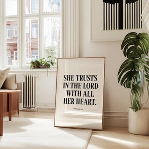 Peut inclure: Une affiche encadrée rectangulaire sur fond beige clair avec un texte noir en police serif indiquant : SHE TRUSTS IN THE LORD WITH ALL HER HEART. PROVERBS 3:5. Le cadre est composé d'un bois fin de couleur marron clair.