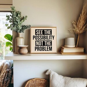 Könnte beinhalten: Ein quadratisches Wandbild mit beigem Hintergrund und fettgedrucktem schwarzem Text: SEE THE POSSIBILITY NOT THE PROBLEM. Das Schild steht auf einem Regal neben einer hohen, strukturierten Vase, einer kleinen Schale und einem Stapel Bücher.