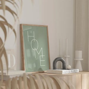 Könnte beinhalten: Ein gerahmter Druck mit dem Wort "HOME" in weißen Buchstaben auf einem salbeigrünen Hintergrund. Der Druck wird auf einem Holzschrank zusammen mit einem dekorativen Bogen, Kerzen und einem Buch ausgestellt.