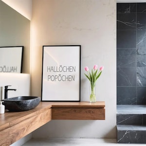 May include: A rectangular framed wall art print with a white background and black text that reads HALLÖCHEN POPÖCHEN. The print is positioned on a wooden vanity next to a clear glass vase containing a bouquet of pink tulips in a bathroom setting.