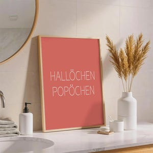 Könnte beinhalten: Ein gerahmter korallenfarbener Druck mit den weißen Worten "HALLÖCHEN POPÖCHEN". Der Druck steht auf einer weißen Arbeitsplatte neben einer weißen Vase mit getrockneten Pflanzen, einem Seifenspender und einem Waschbecken.