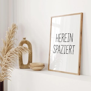 Op de afbeelding: Een rechthoekige ingelijste wandprint met een lichthouten lijst met de zwarte tekst HEREIN SPAZIERT op een witte achtergrond, weergegeven naast een hoog, getextureerd houten decoratief object en een stapel van drie kleine houten kommen.