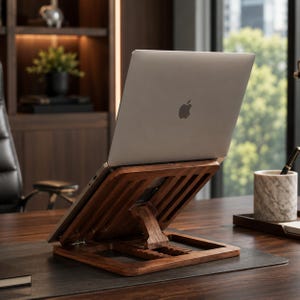 Puede incluir: Un portátil plateado con el logotipo de Apple en la tapa descansa sobre un soporte ajustable de madera marrón oscuro. El soporte presenta un diseño de listones con un brazo de soporte central que permite varios ajustes de altura.