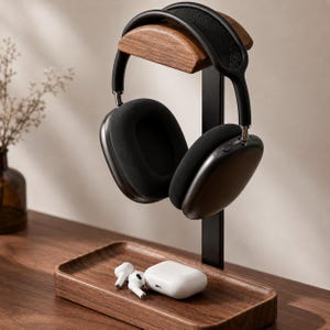May include: A wooden headphone stand with a black metal support, holding black over-ear headphones. A wooden tray at the base holds a white charging case and white earbuds. The stand is on a wooden surface.