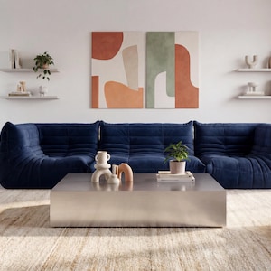 Könnte beinhalten: Ein modernes Wohnzimmer mit einem marineblauen modularen Sofa, einem silbernen Couchtisch und einem gewebten Teppich. Die Wand ist mit abstrakter Kunst und schwebenden Regalen mit Dekoration geschmückt. Eine Topfpflanze sorgt für einen Hauch von Grün.