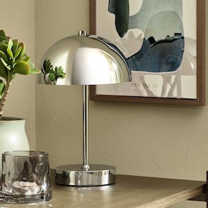 May include: A polished chrome table lamp featuring a smooth, hemispherical dome shade supported by a slender vertical stem and a circular base. The metallic surface reflects the surrounding environment, including a nearby green plant.