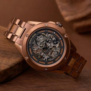 Puede incluir: Un reloj de pulsera de madera marrón con una esfera esqueleto que revela engranajes mecánicos internos. El reloj presenta una caja redonda, una correa de eslabones y el texto DODO DEER impreso en el bisel interior.