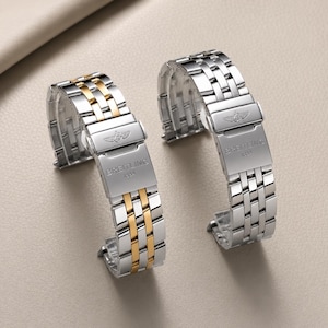Könnte beinhalten: Zwei Metall-Uhrenarmbänder mit Faltschließe. Ein Armband hat ein zweifarbiges Finish in Silber und Gold, das andere ist komplett silberfarben. Beide Schließen sind mit dem Breitling 1884 Logo und dem geflügelten Emblem auf dem Metall graviert.
