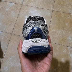 May include: A running shoe with a white sole, silver and blue accents, and black mesh detailing. The shoe is held in a hand, showcasing the heel and rear design. The word "Omni" is visible on the side.