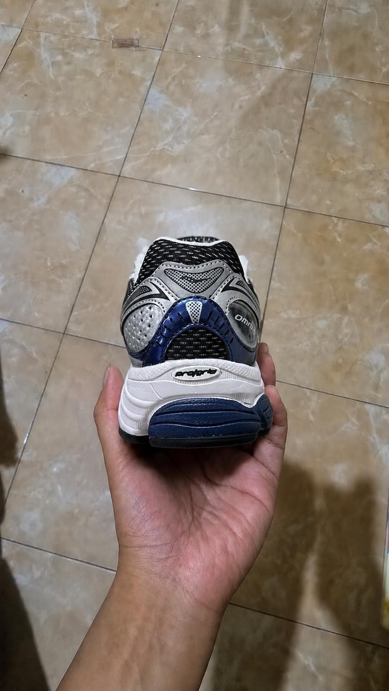 May include: A running shoe with a white sole, silver and blue accents, and black mesh detailing. The shoe is held in a hand, showcasing the heel and rear design. The word "Omni" is visible on the side.