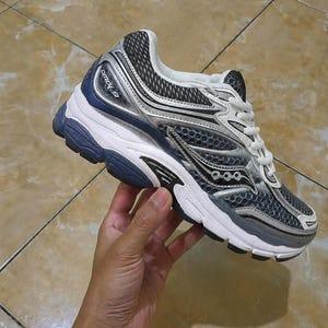 May include: A gray and white athletic shoe with black accents. The shoe features a mesh upper, a white midsole, and a dark blue outsole. The brand name "Omni 9" is visible on the side of the shoe. The shoe is designed for running.