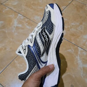 May include: A Saucony running shoe in a sporty design. The shoe features a white, black, and blue color scheme with a mesh upper for breathability. The sole is white and black, providing cushioning and support for running.