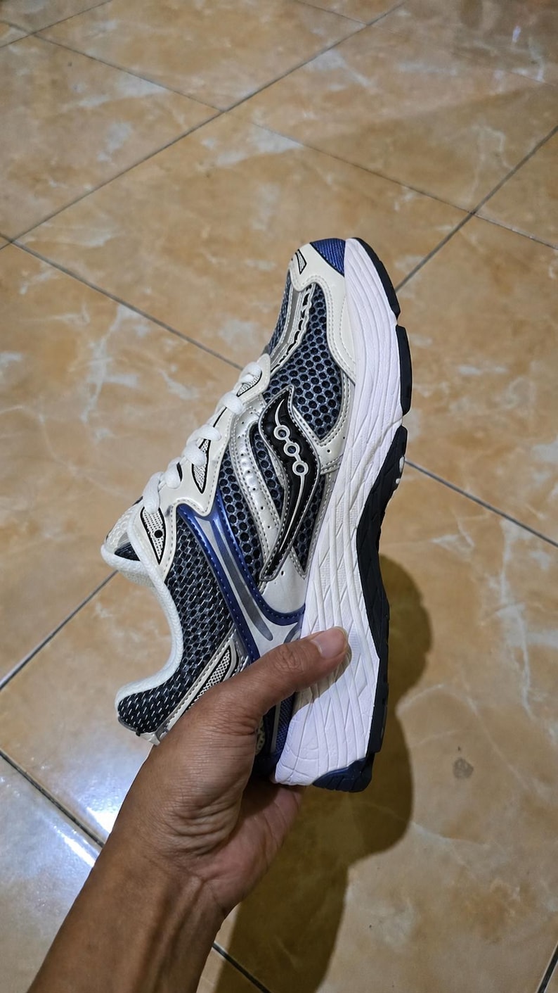 May include: A Saucony running shoe in a sporty design. The shoe features a white, black, and blue color scheme with a mesh upper for breathability. The sole is white and black, providing cushioning and support for running.