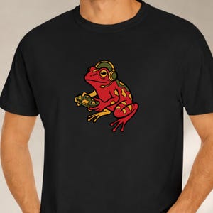 Op de afbeelding: Een zwart t-shirt met een grafische illustratie van een rode kikker met gele vlekken. De kikker draagt een olijfgroene koptelefoon met een microfoon en houdt met beide handen een groene videogamecontroller vast in een zittende houding.