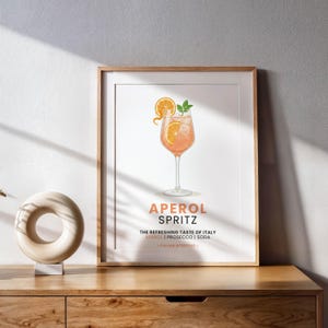 May include: A framed print featuring an Aperol Spritz cocktail illustration. The drink is in a tall glass, garnished with orange slices and mint. Text at the bottom reads "APEROL SPRITZ" and "THE REFRESHING TASTE OF ITALY". The print is in a wooden frame.